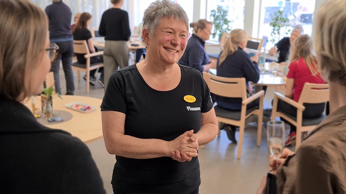 En dam som står och ler samtidigt som det sitter flera runt om henne på stolar och samtalar.