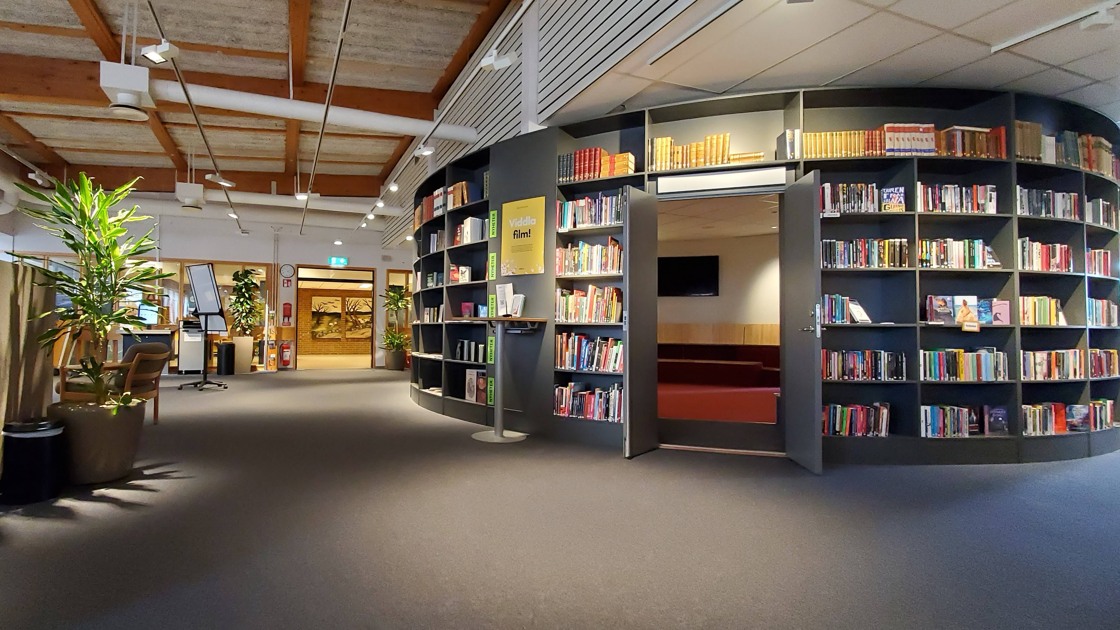 Miljöbild inifrån Tibro bibliotek. Stort öppet rum med mörk matta. Till höger i bild rotunda med bokhyllor och böcker.