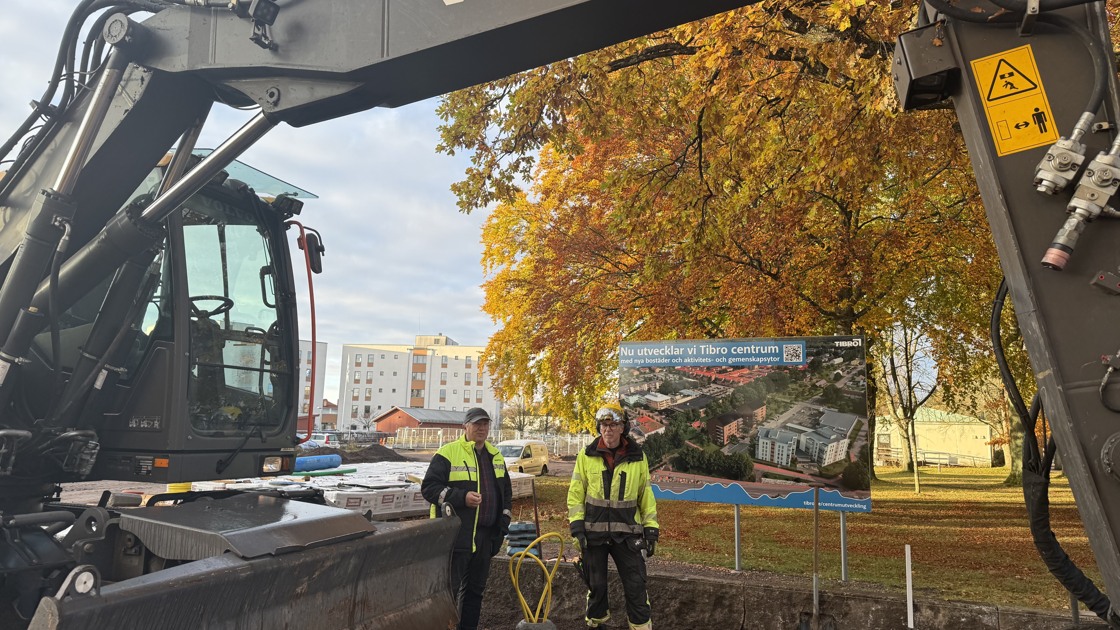 Två byggarbetare står framför en skylt med texten Nu utvecklar vi Tibro centrum. Det är en grävmaskin framför personerna.