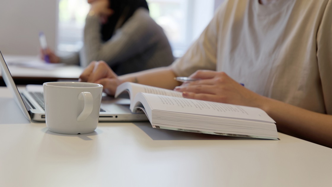 En elevs händer när hon läser studielitteratur framför en laptop med en vit mugg bredvid på bordet.