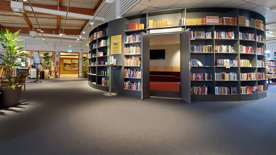 Miljöbild inifrån Tibro bibliotek. Stort öppet rum med mörk matta. Till höger i bild rotunda med bokhyllor och böcker.