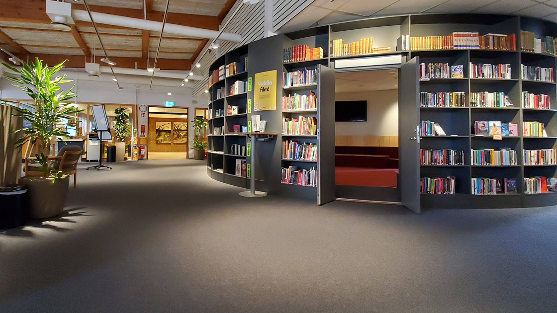 Miljöbild inifrån Tibro bibliotek. Stort öppet rum med mörk matta. Till höger i bild rotunda med bokhyllor och böcker.