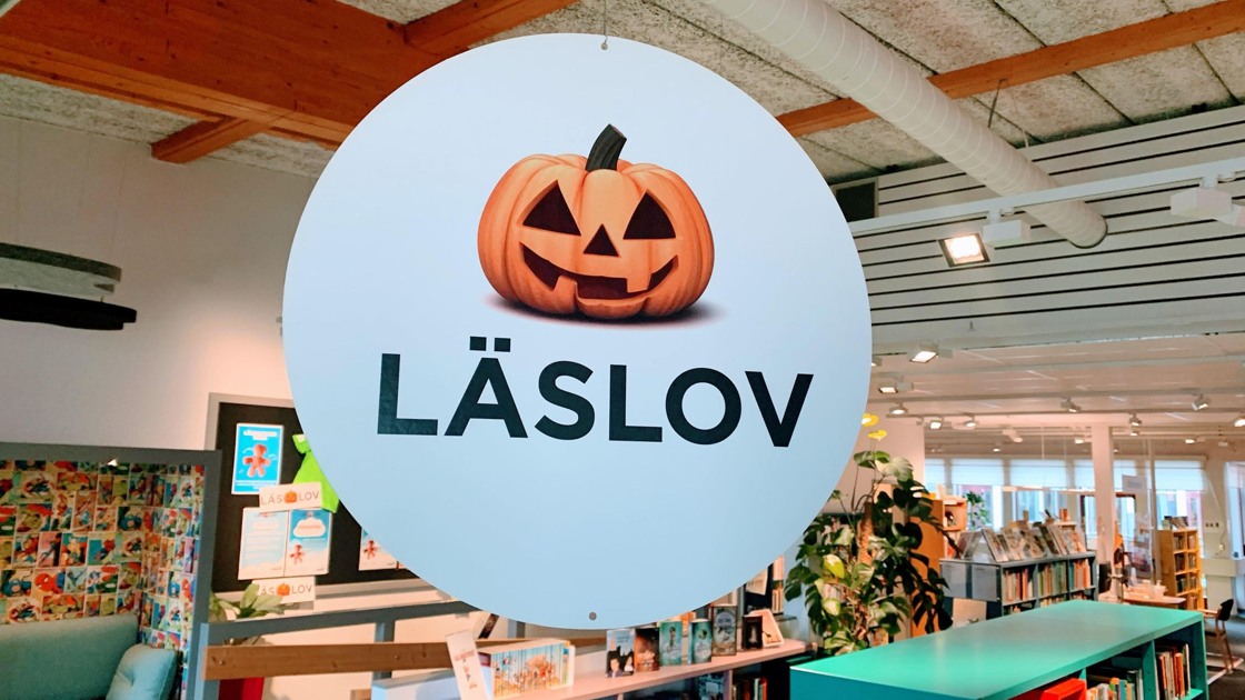 Hängande rund vit skylt med pumpa och text Läslov, I bakgrunden bokhyllor.