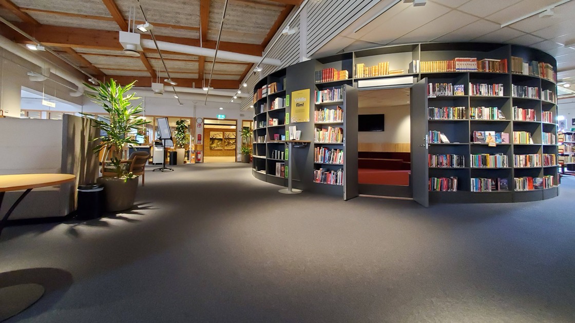 Miljöbild inifrån Tibro bibliotek. Stort öppet rum med mörk matta. Till höger i bild rotunda med bokhyllor och böcker.
