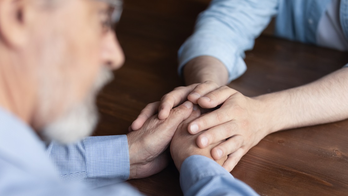 Supportive hands. Close up caring young man adult child caregiver sit at table facing old man retiree support comfort express empathy. Geriatric psychologist talk to elder patient on therapy session