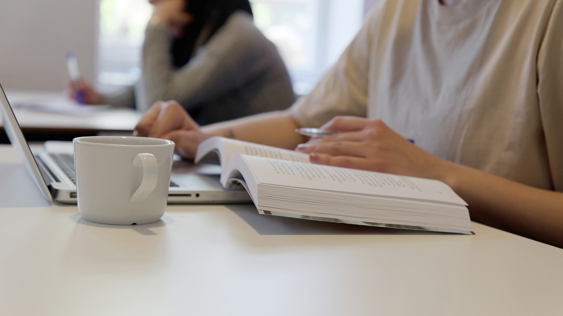 En elevs händer när hon läser studielitteratur framför en laptop med en vit mugg bredvid på bordet.
